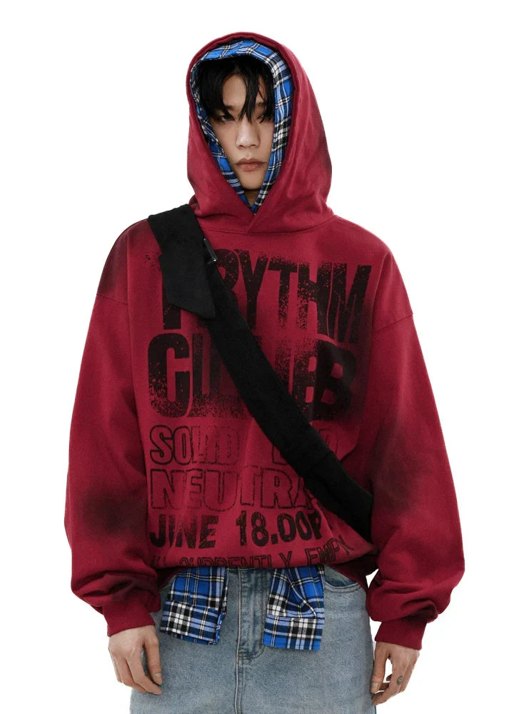 黒い文字が入ったキャンバス パーカーを着て、その下に青いチェック柄のフードをかぶり、黒いクロスボディ バッグを持った人物が、薄いデニム ジーンズをはき、白い背景に立ってカメラを見ています。.