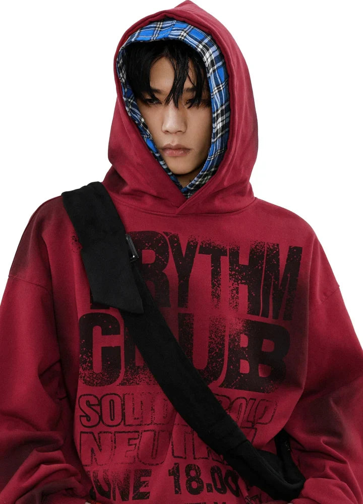 赤い文字と黒い文字が入ったキャンバス パーカーを着て、青いチェック柄のフード付きシャツを着て、下を向いている人物。白い背景に、黒いクロスボディ バッグを肩にかけています。.