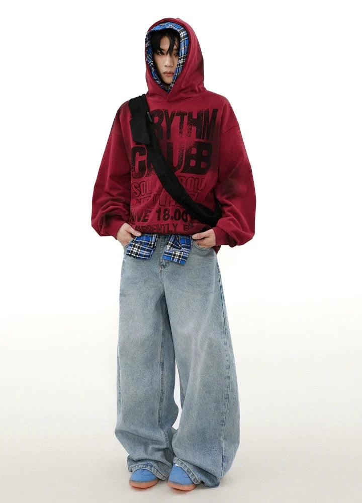 黒のグラフィックがあしらわれた赤の CANVAS HOODIE を着用し、ゆったりとしたライトブルーのジーンズ、その下にブルーのチェック柄シャツ、ライトブルーとピンクの靴を合わせています。アクセサリーは、無地の白い背景に黒のクロスボディバッグです。.