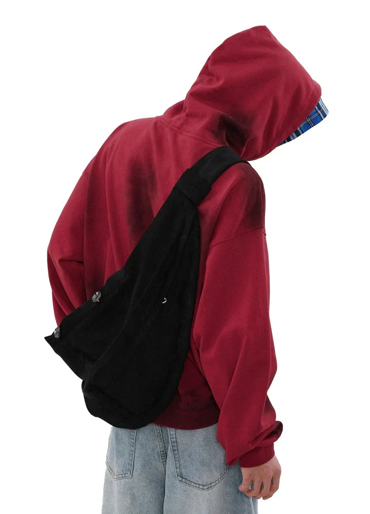 赤いフードをかぶり、チェック柄の裏地が見えるキャンバス パーカーを着た人物が、カメラから背を向けています。人物はライトブルーのジーンズと黒いショルダー バッグを身に着けており、無地の白い背景にいます。.